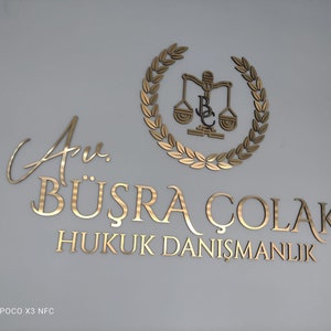 May include: A gold-colored wall sign featuring the text Av. B&Uuml;ŞRA &Ccedil;OLAK HUKUK DANIŞMANLIK in a serif font, accompanied by a circular laurel wreath emblem surrounding a scale of justice icon with the letters BC integrated into the center design.