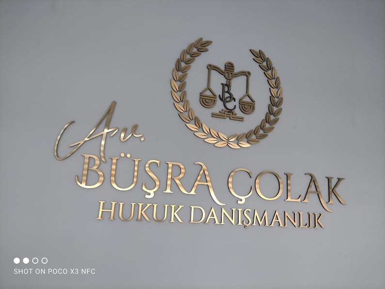 May include: A gold-colored wall sign featuring the text Av. B&Uuml;ŞRA &Ccedil;OLAK HUKUK DANIŞMANLIK in a serif font, accompanied by a circular laurel wreath emblem surrounding a scale of justice icon with the letters BC integrated into the center design.