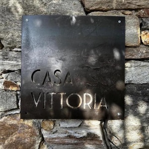 May include: A dark, square metal sign with the words "CASA VITTORIA" cut out. The sign is mounted on a stone wall. The letters are in a simple, sans-serif font. The sign is held in place by four screws.