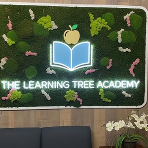May include: A rectangular wall sign featuring a green moss background with scattered pink and white floral accents. Centered is a light blue illuminated open book icon topped with a yellow apple, and the text THE LEARNING TREE ACADEMY in white.