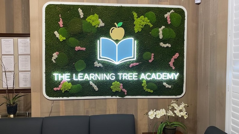 May include: A rectangular wall sign featuring a green moss background with scattered pink and white floral accents. Centered is a light blue illuminated open book icon topped with a yellow apple, and the text THE LEARNING TREE ACADEMY in white.