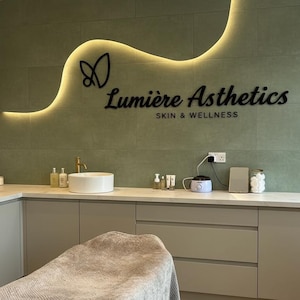 May include: A treatment room featuring a sage green tiled wall with a backlit wavy accent and the black text Lumi&egrave;re Asthetics SKIN & WELLNESS. The room includes a white countertop, a round sink, a gray treatment bed, and various skincare bottles.