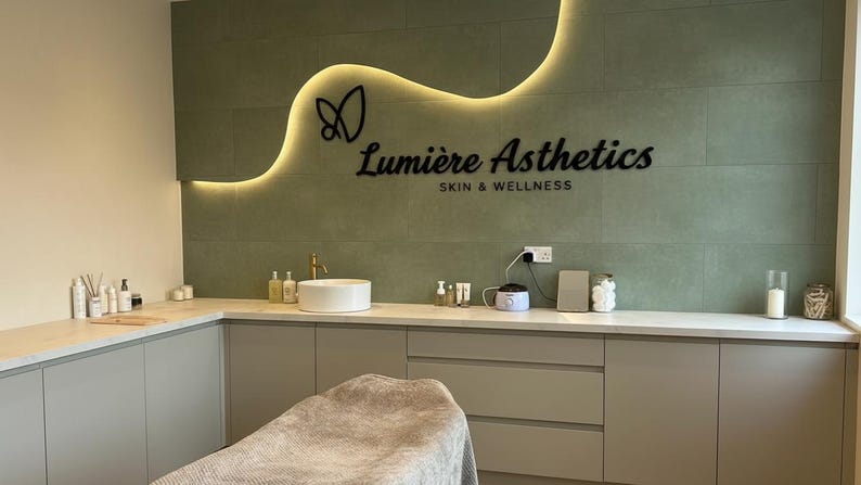 May include: A treatment room featuring a sage green tiled wall with a backlit wavy accent and the black text Lumi&egrave;re Asthetics SKIN & WELLNESS. The room includes a white countertop, a round sink, a gray treatment bed, and various skincare bottles.
