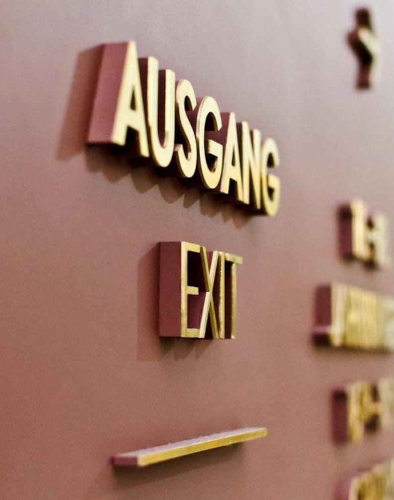 K&ouml;nnte beinhalten: Nahaufnahme von Wandzeichen mit goldfarbenen Buchstaben. Das Wort "AUSGANG" ist sichtbar, zusammen mit dem Wort "EXIT". Die Schilder sind an einer mauvefarbenen Wand angebracht. Weitere Schilder sind im Hintergrund zu sehen.