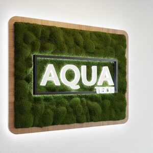 May include: A rectangular wall sign featuring a thick layer of green preserved moss framed in light wood. Centered is a black rectangular frame containing the white illuminated text AQUA TECH, with a soft glow emanating from behind the sign.
