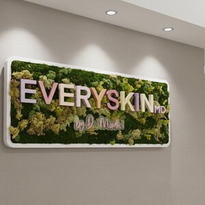 May include: A rectangular wall sign featuring a white frame filled with preserved green moss. The sign displays the text EVERYSKIN MD in pastel-colored 3D letters, with the script text by D. Madhi positioned directly underneath the main lettering.