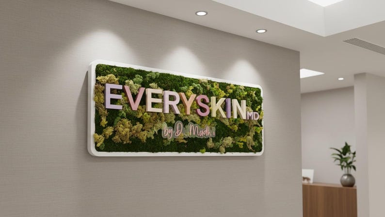 May include: A rectangular wall sign featuring a white frame filled with preserved green moss. The sign displays the text EVERYSKIN MD in pastel-colored 3D letters, with the script text by D. Madhi positioned directly underneath the main lettering.