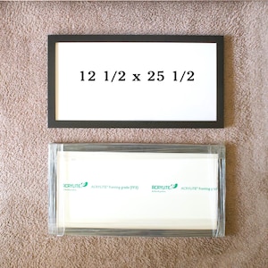 May include: A rectangular black picture frame with white matting displaying the text 12 1/2 x 25 1/2, positioned above a second rectangular frame wrapped in clear plastic with a white protective sheet labeled Acrylite framing grade FF3.