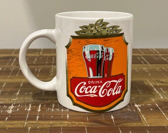 May include: White ceramic mug with a Coca-Cola logo and two glasses of Coca-Cola. The logo is red and gold with the words "Drink Coca-Cola" in a white banner.