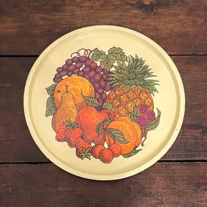 May include: A round, cream-colored metal serving tray with a colorful fruit design. The tray features a pineapple, grapes, cherries, strawberries, a pear, and oranges.