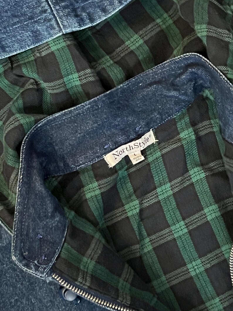 May include: Close-up of a denim jacket with a green and black plaid lining. The jacket has a zipper and a collar. The tag reads "NorthStyle". The denim is a medium wash blue.