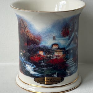 May include: A white ceramic mug with a gold rim and base. The mug features a scenic painting of a cottage at dusk, with a waterfall and swans. The text "Evening at Swanbrooke Cottage" is displayed on the front.