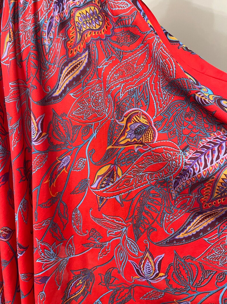 May include: Red fabric with a paisley pattern. The pattern features intricate floral designs in shades of blue, purple, yellow, and orange.