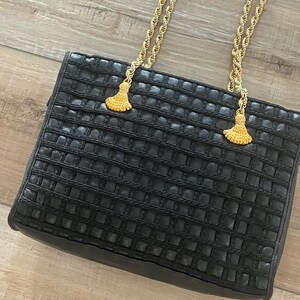 May include: A black handbag featuring a textured grid pattern across the front surface. The bag includes two gold-tone twisted rope shoulder straps that are attached to the top with decorative gold-tone tassels at each connection point.