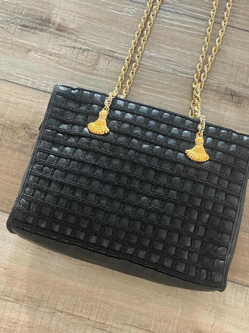 May include: A black handbag featuring a textured grid pattern across the front surface. The bag includes two gold-tone twisted rope shoulder straps that are attached to the top with decorative gold-tone tassels at each connection point.