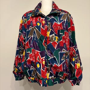 May include: A long-sleeve jacket featuring a vibrant, multi-colored abstract geometric pattern in shades of red, navy blue, purple, green, and yellow. The jacket has a high collar, a front zipper closure, and elasticized cuffs and hemline.