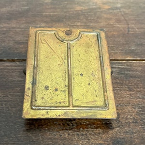 May include: A rectangular, antique brass-colored metal clip with a decorative design. The clip has a small hole at the top and a hinged clasp on the back. The surface shows signs of age and wear.