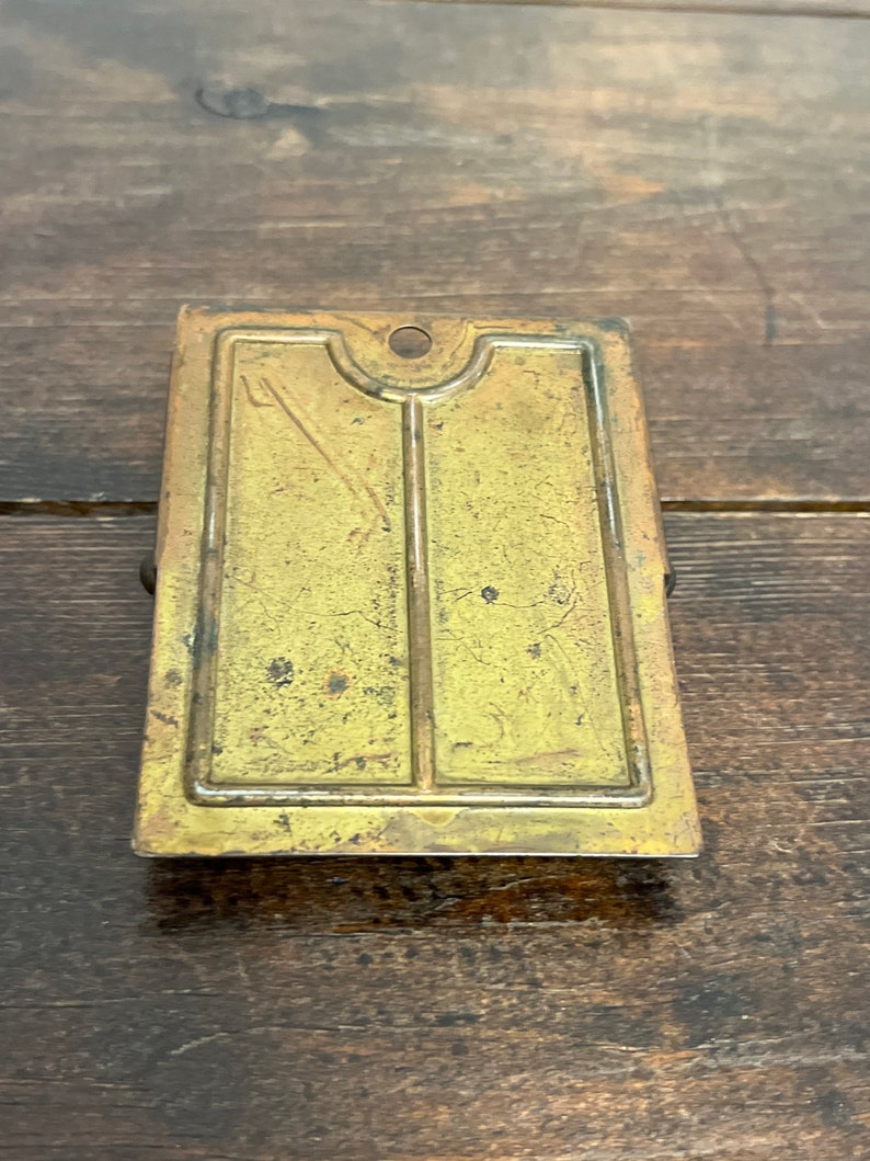 May include: A rectangular, antique brass-colored metal clip with a decorative design. The clip has a small hole at the top and a hinged clasp on the back. The surface shows signs of age and wear.