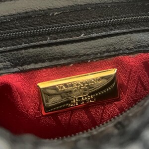 May include: A black bag interior featuring a red fabric lining with a repeating V pattern. A rectangular gold-tone metal plaque is centered, engraved with the text VALENTINO, a V logo, and the words MADE IN ITALY. A black zipper is visible above.