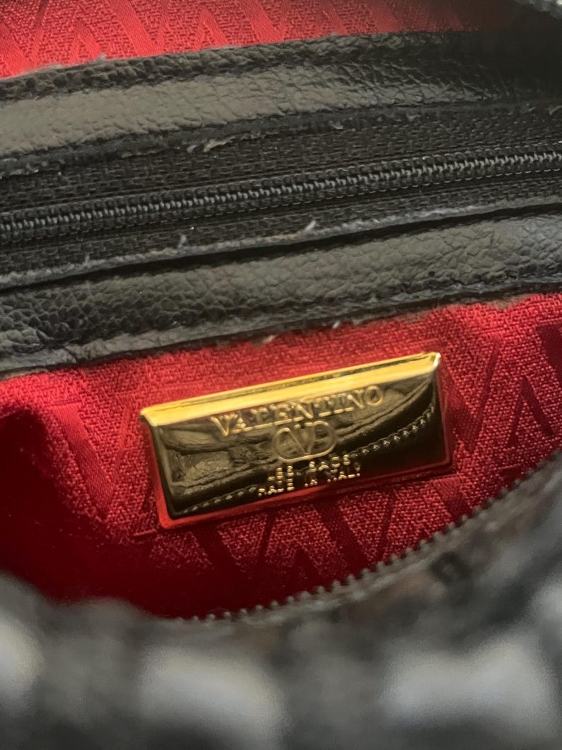 May include: A black bag interior featuring a red fabric lining with a repeating V pattern. A rectangular gold-tone metal plaque is centered, engraved with the text VALENTINO, a V logo, and the words MADE IN ITALY. A black zipper is visible above.