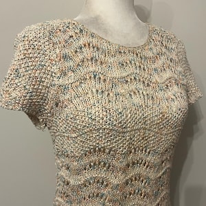 May include: A short-sleeved, cream-colored knit top with a round neckline. The top features a textured knit pattern with subtle flecks of blue, peach, and brown. Scalloped detailing adds a decorative element to the design. The garment is displayed on a mannequin.