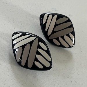 May include: A pair of black earrings with a leaf-like shape. Each earring features a pattern of white and gray oblong shapes. The earrings have a glossy finish and are displayed on a white surface.