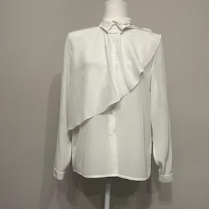 May include: A white long-sleeved blouse with a ruffled collar and a single button closure. The blouse is on a white mannequin.