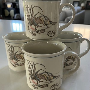 May include: A set of four off-white ceramic mugs featuring a detailed illustration of seashells, sand dollars, and beach grass. Each mug has a smooth handle and a thin blue line encircling the upper rim, showcasing a consistent coastal design.