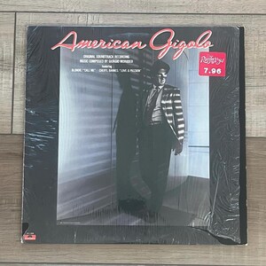 May include: Sealed vinyl record for the "American Gigolo" soundtrack. The album cover features the title in pink script, a man in a striped suit, and the text "ORIGINAL SOUNDTRACK RECORDING" and "MUSIC COMPOSED BY GIORGIO MORODER".