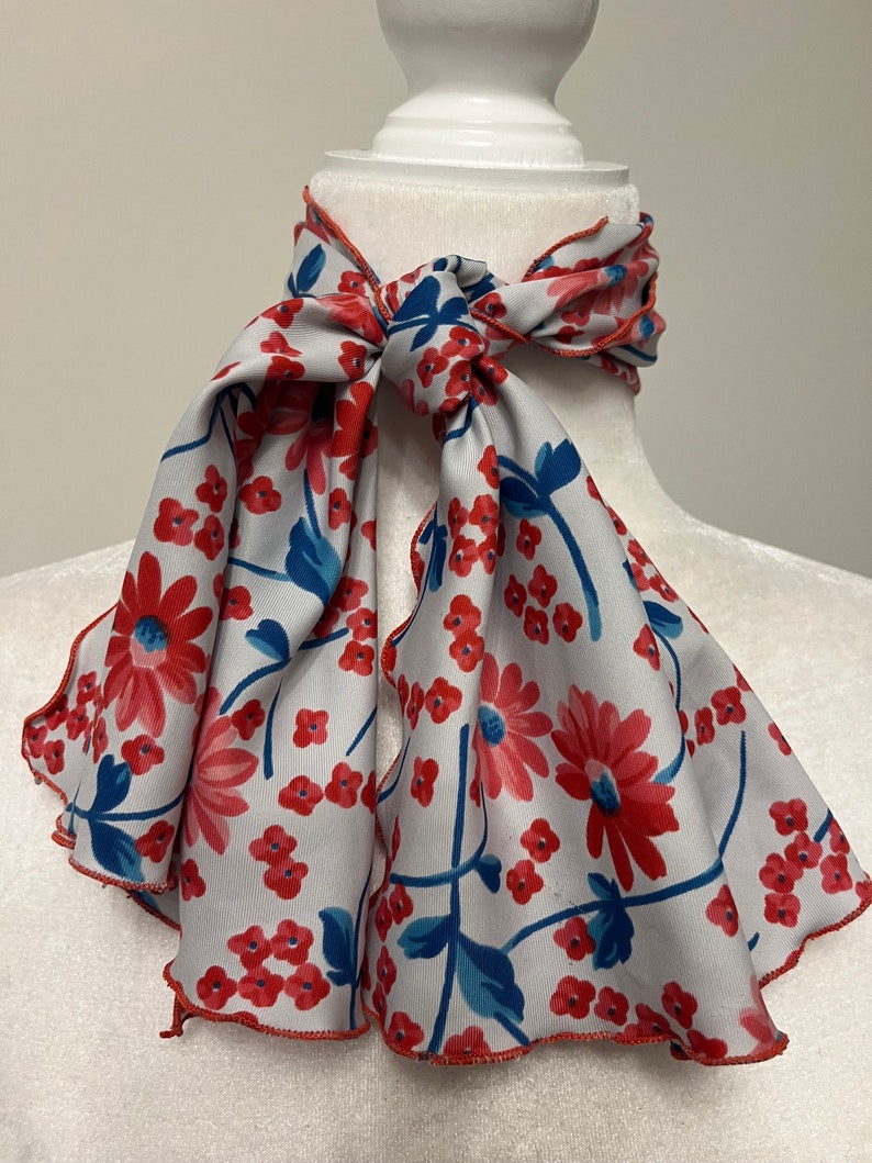 May include: A white scarf with a red and blue floral pattern. The scarf has a scalloped edge and is tied in a bow around a white mannequin.