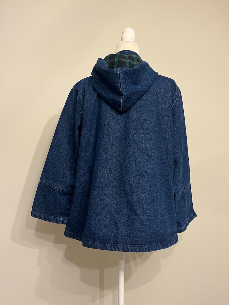 May include: A dark blue denim jacket with a hood. The hood lining features a green and black plaid pattern. The jacket has a relaxed fit and wide sleeves, displayed on a white mannequin.