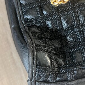 May include: A black handbag featuring a textured, grid-like pattern created by woven cord detailing across the surface. The bag includes a thick, polished gold-tone metal chain link strap attached to the top edge, showcasing a metallic finish.
