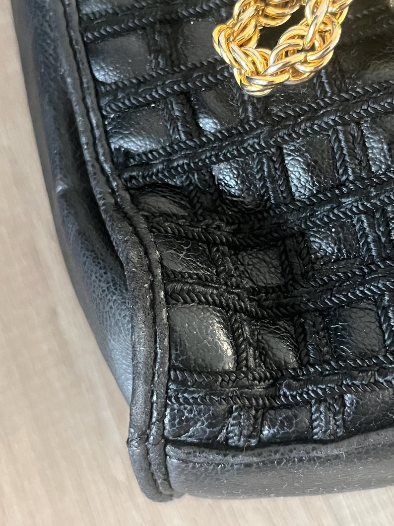 May include: A black handbag featuring a textured, grid-like pattern created by woven cord detailing across the surface. The bag includes a thick, polished gold-tone metal chain link strap attached to the top edge, showcasing a metallic finish.