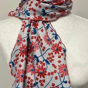 May include: A white scarf with a red and blue floral pattern. The scarf has a red border and is made of a soft, lightweight fabric.