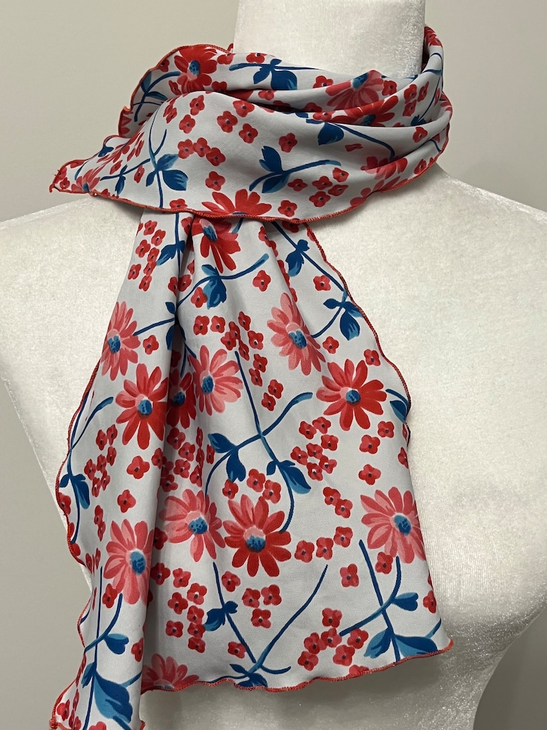 May include: A white scarf with a red and blue floral pattern. The scarf has a red border and is made of a soft, lightweight fabric.