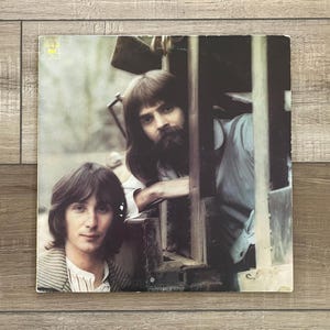 May include: Vintage vinyl record album cover featuring two men. One man has long hair and a beard, leaning out of a wooden structure. The other man has short hair and is wearing a striped shirt. The album cover has a faded, vintage aesthetic.
