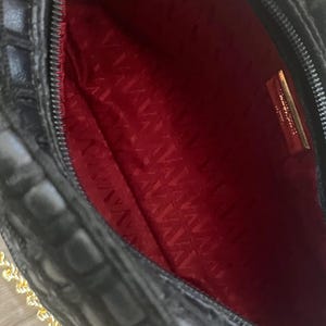 May include: A black handbag with a textured exterior and a vibrant red interior lining. The bag features a gold-toned metal chain strap and a rectangular gold-toned metal plaque attached to the inner lining with visible text.