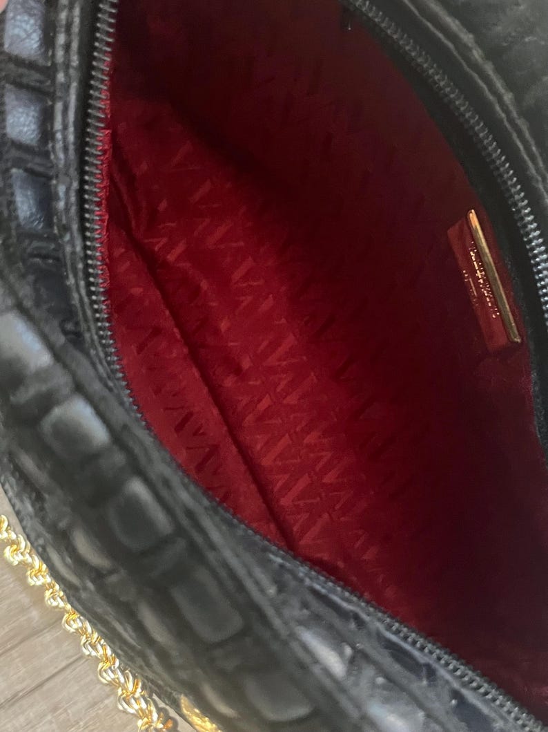 May include: A black handbag with a textured exterior and a vibrant red interior lining. The bag features a gold-toned metal chain strap and a rectangular gold-toned metal plaque attached to the inner lining with visible text.