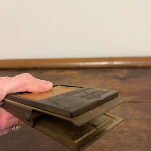 May include: Vintage metal clip with a rectangular shape. The clip has a hinged design with a top section that reads "The American Clip Company". The clip is made of metal with a brass and dark metal finish.