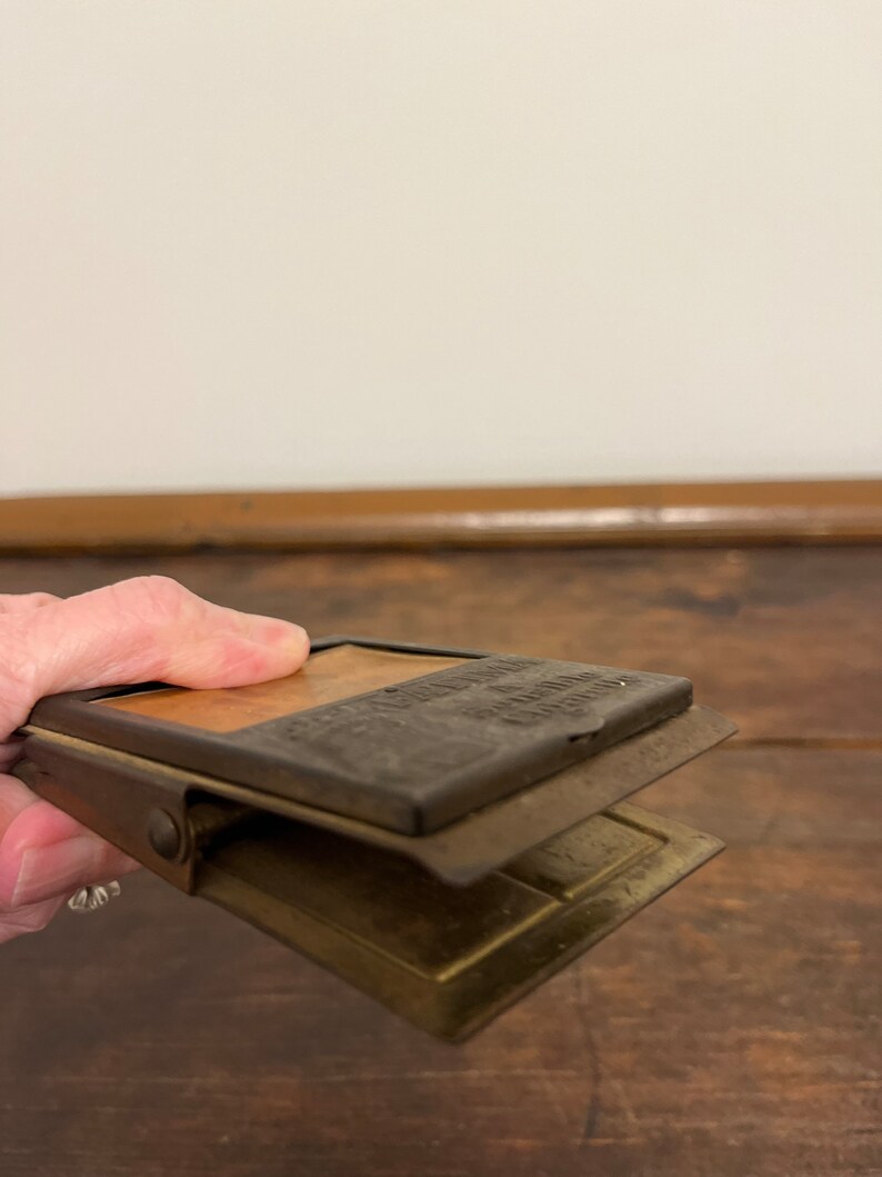 May include: Vintage metal clip with a rectangular shape. The clip has a hinged design with a top section that reads "The American Clip Company". The clip is made of metal with a brass and dark metal finish.