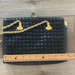 May include: A rectangular black handbag with a woven grid texture, featuring a gold-tone rope chain strap with two decorative tassels. A wooden ruler placed across the front shows the bag measures approximately 10.5 inches in width.
