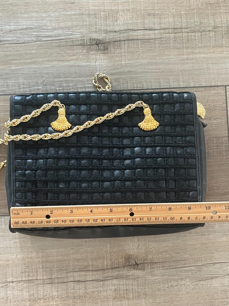 May include: A rectangular black handbag with a woven grid texture, featuring a gold-tone rope chain strap with two decorative tassels. A wooden ruler placed across the front shows the bag measures approximately 10.5 inches in width.