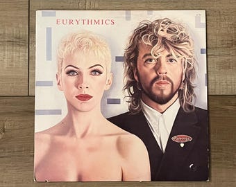 Può includere: Copertina dell'album degli Eurythmics con una donna bionda con rossetto rosso e un uomo in abito nero. Il titolo dell'album, "Eurythmics", è stampato in rosso in alto. L'uomo ha capelli lunghi e ricci e la barba.