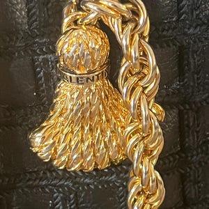 May include: A gold-tone metal tassel charm featuring a textured, fan-shaped design with a rounded top. The word VALENTINO is engraved on a smooth band near the top. The charm is attached to a thick, twisted gold-tone metal chain link.