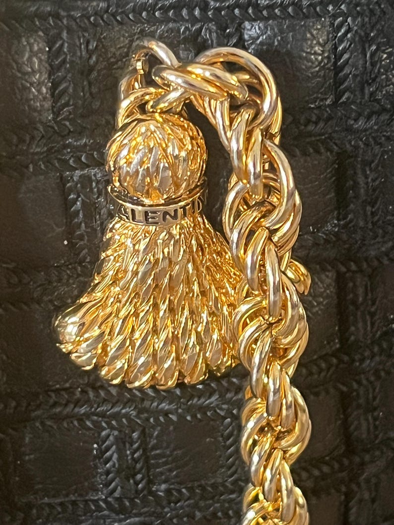 May include: A gold-tone metal tassel charm featuring a textured, fan-shaped design with a rounded top. The word VALENTINO is engraved on a smooth band near the top. The charm is attached to a thick, twisted gold-tone metal chain link.