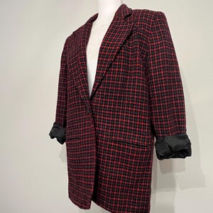 Puede incluir: Un blazer a cuadros rojos y negros sobre un maniquí blanco. El blazer tiene un cuello con solapas clásico, mangas largas con puños negros y un corte entallado. La tela tiene una textura tejida.
