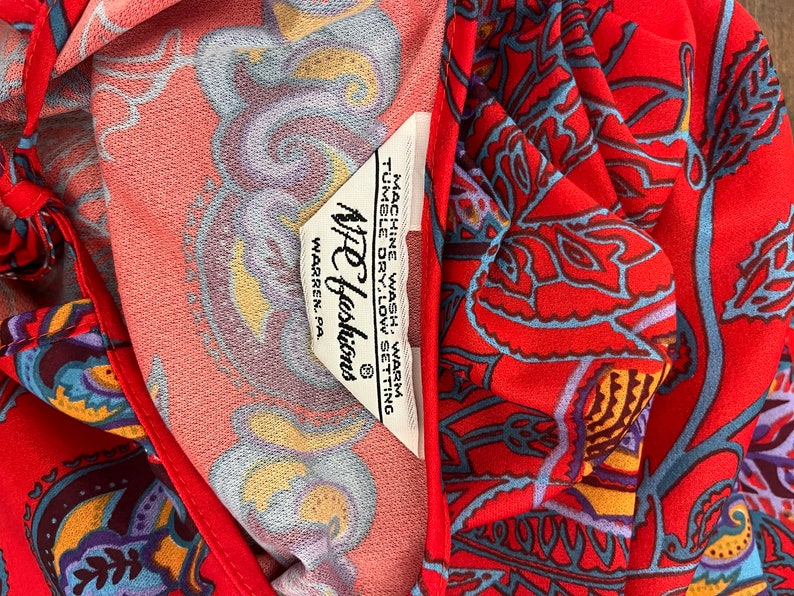 May include: A red fabric with a paisley pattern. The fabric has a white tag with the text "NFC Fashions" and "WARREN, PA.". The tag also includes instructions for washing and drying the fabric.