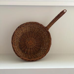 May include: A round, brown woven wicker basket with a single long, sturdy handle extending from one side. The basket features a circular base with a tight, intricate weave pattern that expands into an open, airy lattice design toward the rim.