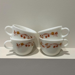 May include: Three white milk glass cups with rounded handles and a repeating orange and brown floral pattern featuring small blossoms and wheat stalks. The cups are arranged with two stacked on the left and one positioned to the right.