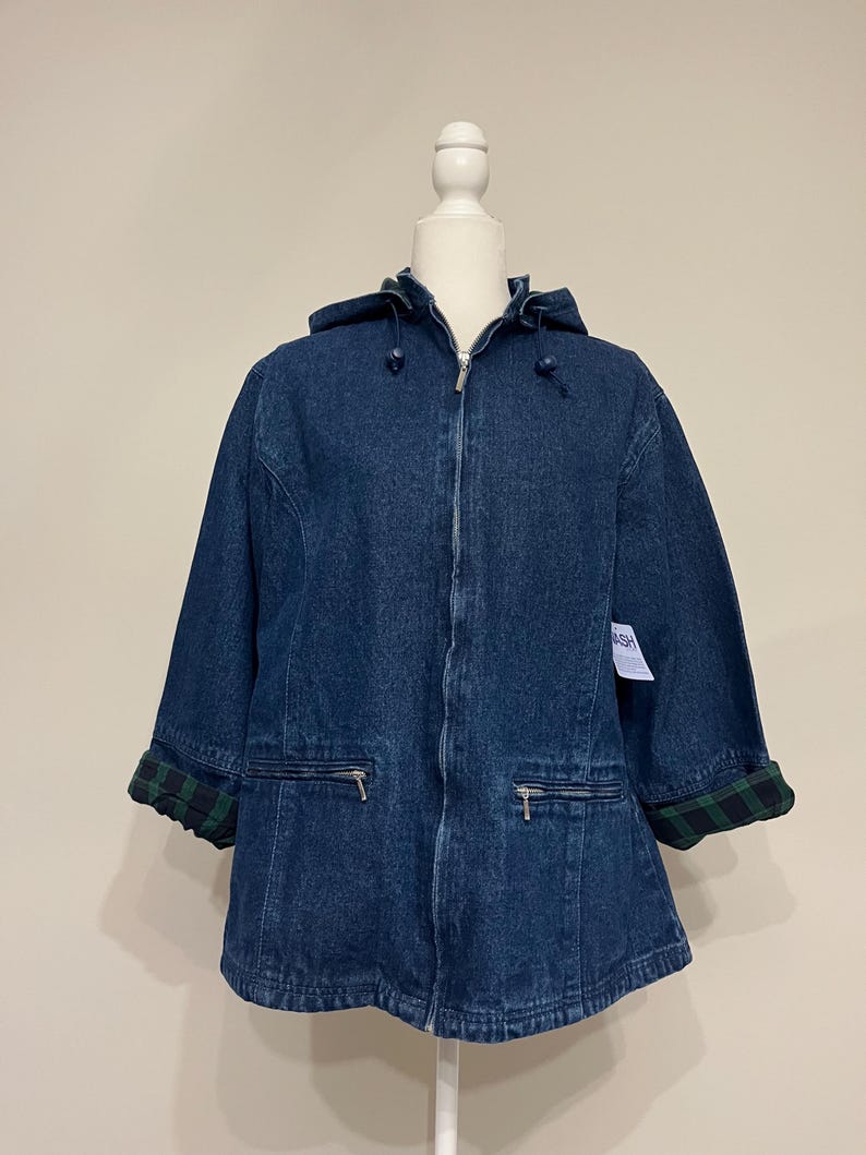 May include: A dark blue denim jacket with a hood and a zipper closure. The jacket has two front pockets with zippers and green and black plaid cuffs. The jacket is displayed on a white mannequin.